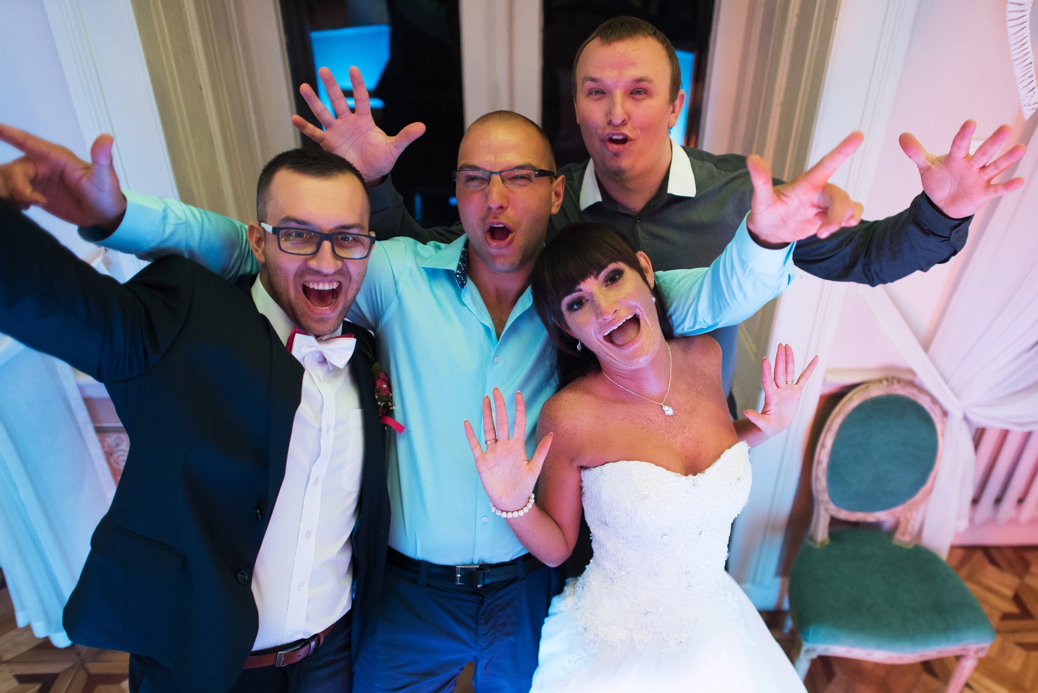 Radosna grupa weselna pozuje do zdjęcia: panna młoda w sukni, panowie w koszulach i marynarce, wszyscy z szerokimi uśmiechami i uniesionymi rękami. Tło stanowią białe zasłony i krzesło.