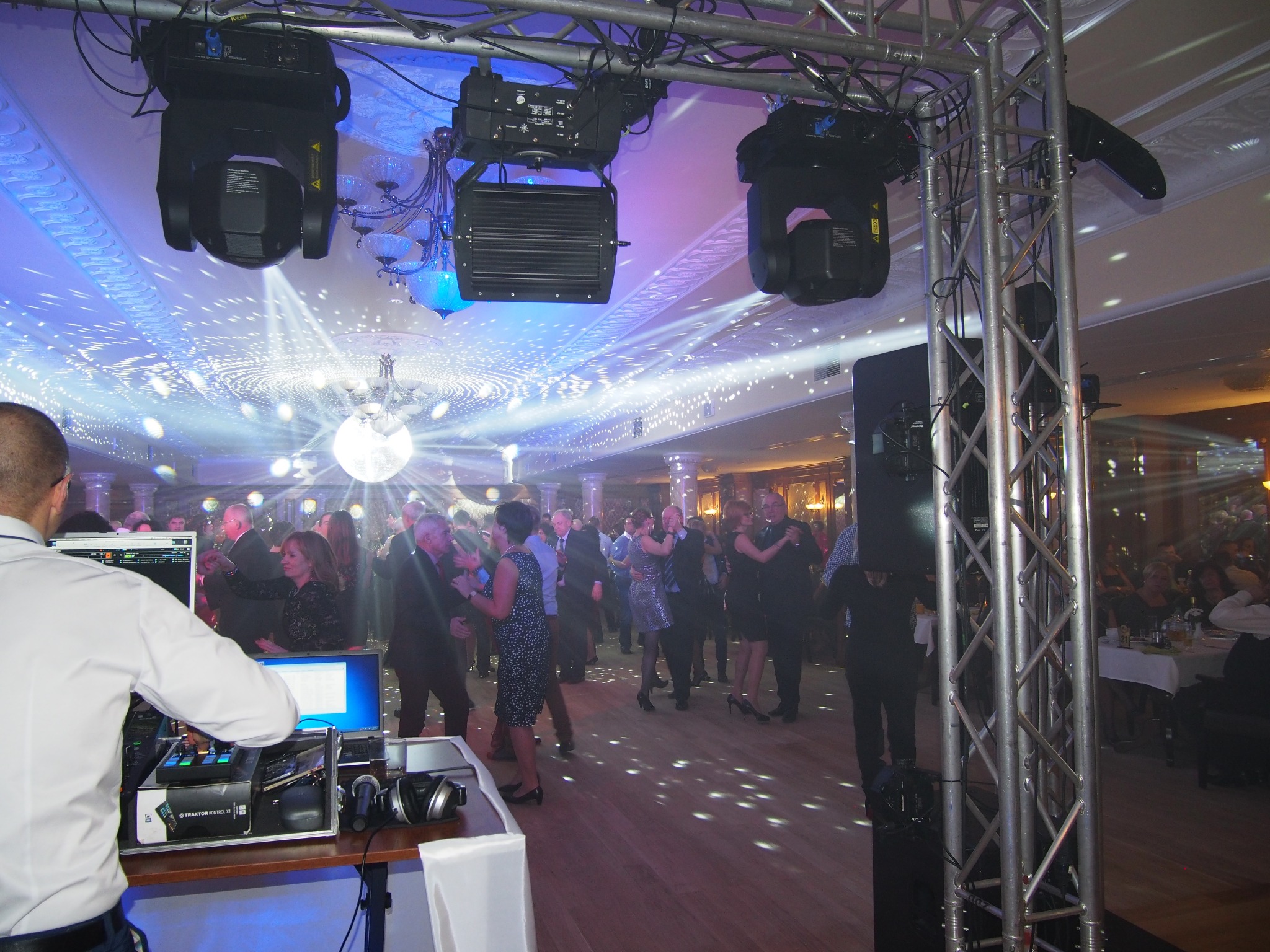 Parkiet w blasku świateł dyskotekowych, DJ za konsoletą, bawiący się ludzie w tle. Oświetlenie sceniczne zawieszone na konstrukcji kratownicowej.