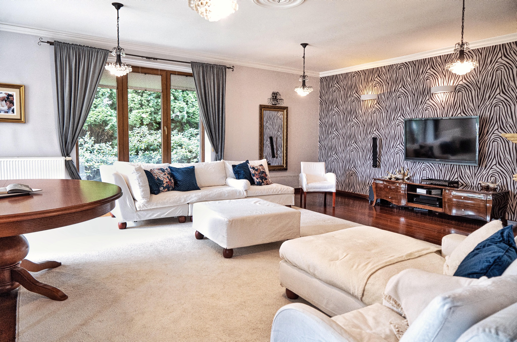 Elegancki salon z białymi sofami, wzorzystą tapetą i drewnianymi meblami. Okno z widokiem na zieleń. Lampa wisząca nad stołem. Lustro w złotej ramie.