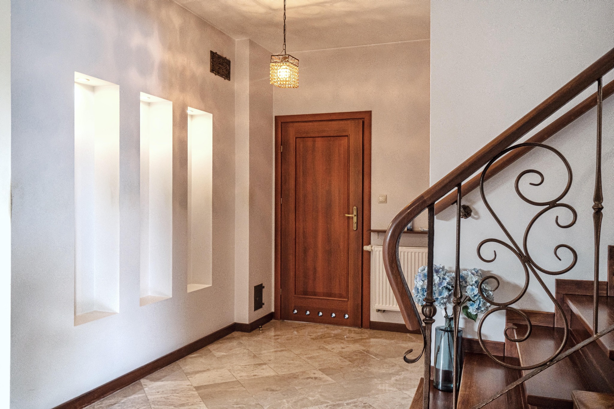 Korytarz z drewnianymi schodami i balustradą, dekoracyjnymi wnękami w ścianie, oświetleniem i marmurową podłogą. Widok na drzwi wejściowe i grzejnik.