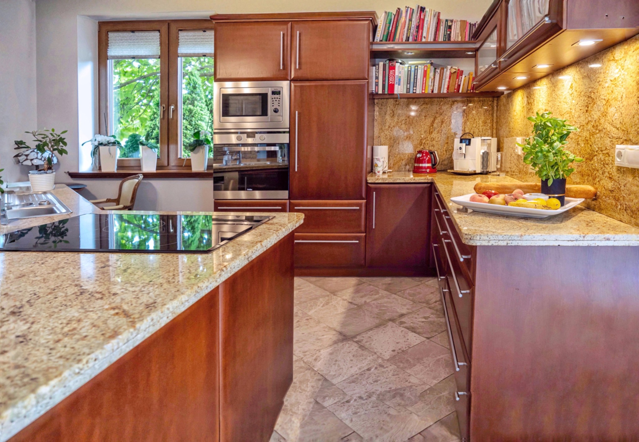 Nowoczesna kuchnia z drewnianymi szafkami, granitowym blatem i wyspą. Widoczne owoce na tacy, okno z zielenią i książki na półkach. Ciepłe oświetlenie.