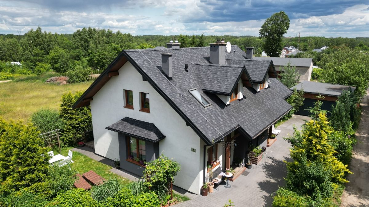 Dwupiętrowy dom z ciemnoszarym dachem wielospadowym, białymi ścianami i brązowymi oknami, otoczony zielenią. Widok z góry na zadbany ogród i podjazd.