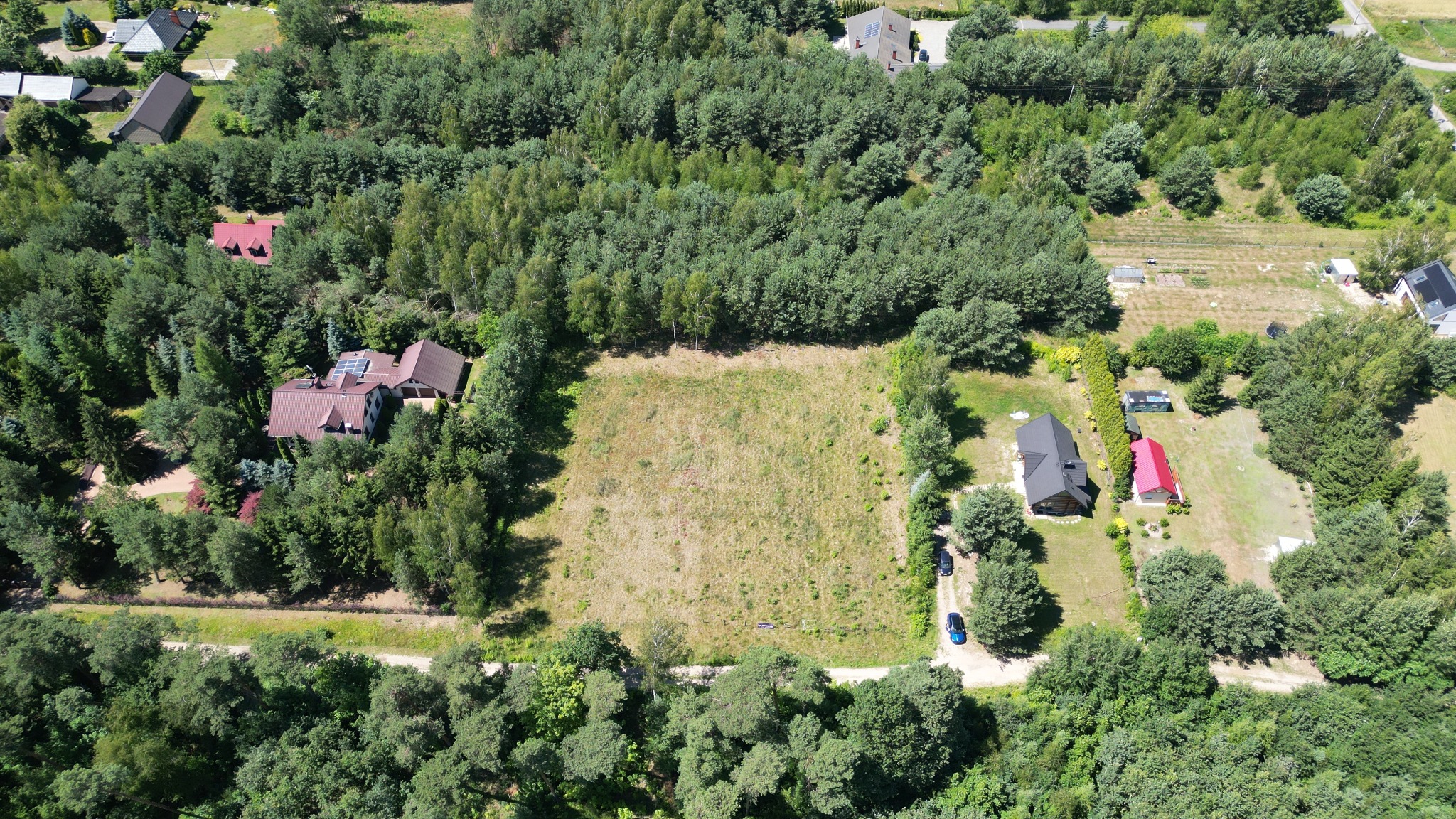 Zdjęcie z drona przedstawia działkę budowlaną otoczoną lasem i zabudowaniami. Widoczny teren przygotowany pod inwestycję, z dostępem do drogi i bliskością natury.