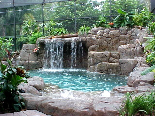 Kaskadowy wodospad z kamieni wpadający do turkusowej wody, otoczony bujną roślinnością i zabezpieczony siatką, widok z poziomu ziemi.