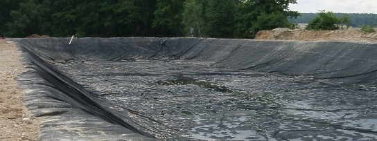 Wyłożony czarną folią zbiornik retencyjny na wodę, częściowo wypełniony, z ziemnym brzegiem i widocznym lasem w tle.