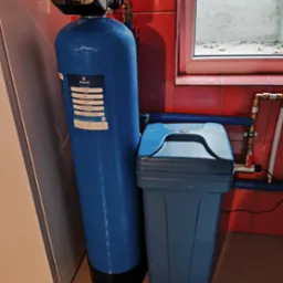 Błękitny filtr wody Klarwod 12x52 z szarą obudową na tle czerwonych płytek i okna. Widoczne rury instalacji wodnej. Filtr gotowy do użytku.