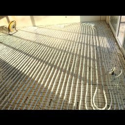 TYRKA INSTALACJE - MARIUSZ TYRKA - Białe rurki ogrzewania podłogowego ułożone na siatce, z widocznym żółtym wentylatorem i fragmentem okna, oświetlone naturalnym światłem.