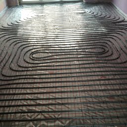 TYRKA INSTALACJE - MARIUSZ TYRKA - Ułożone rury ogrzewania podłogowego na folii refleksyjnej z czerwoną kratką, widok z góry, równoległe ułożenie rur w kształcie pętli, jasne światło z okna.