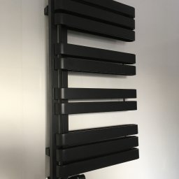 TYRKA INSTALACJE - MARIUSZ TYRKA - Czarny, designerski grzejnik łazienkowy, zamontowany na ścianie z białych płytek. Widoczny zawór termostatyczny. Minimalistyczny styl.