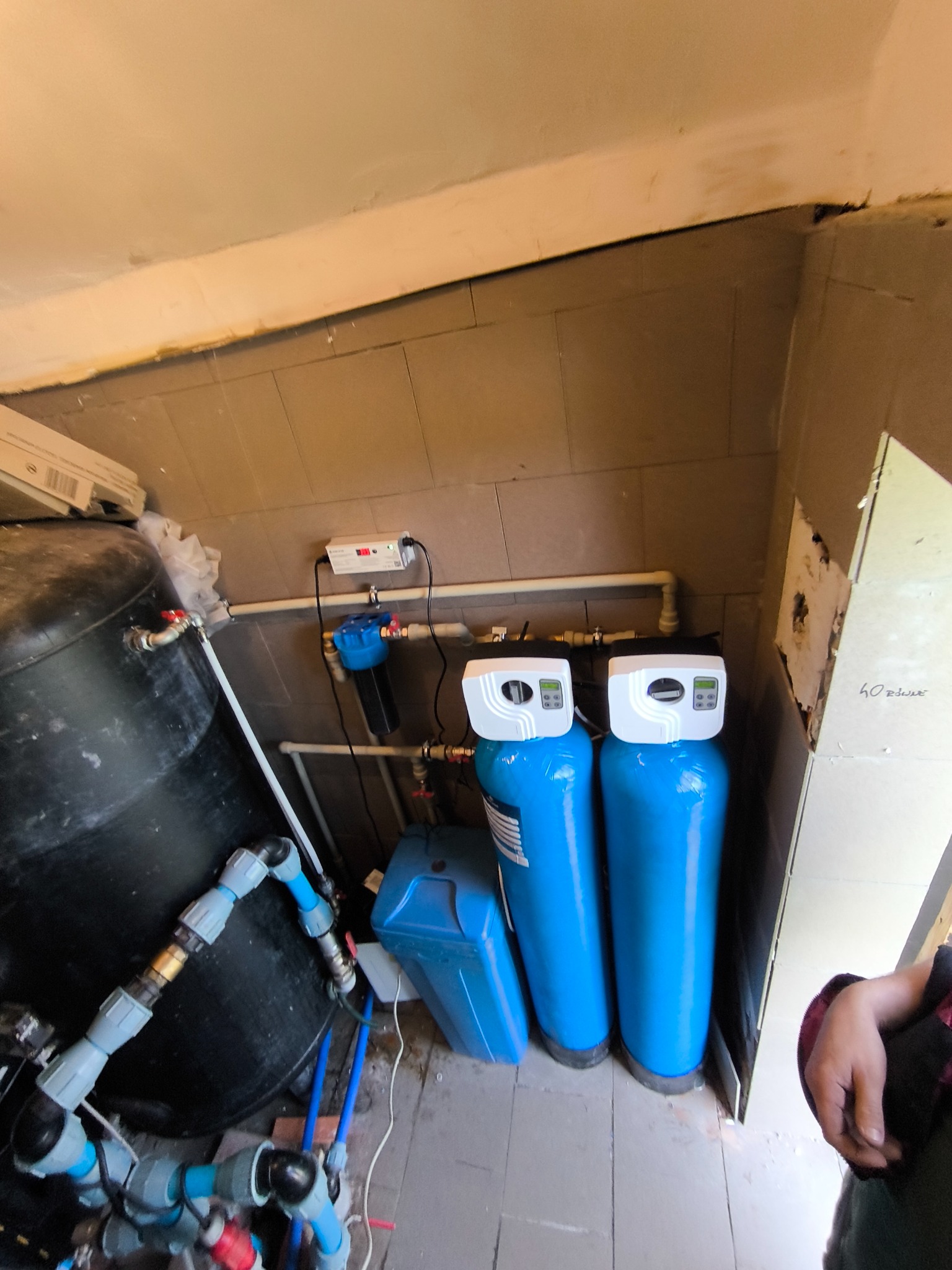 Zmontowany system uzdatniania wody z dwoma niebieskimi zbiornikami, czarnym hydroforem i instalacją hydrauliczną w pomieszczeniu z brązowymi płytkami.