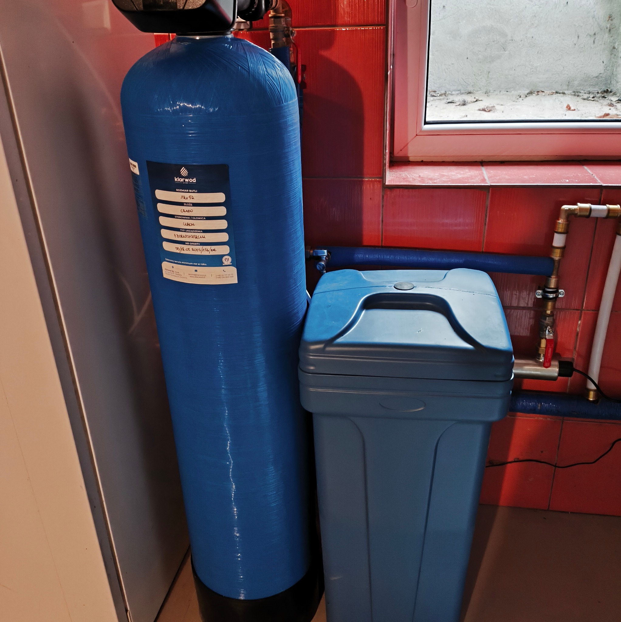 Błękitny filtr wody Klarwod 12x52 z szarą obudową na tle czerwonych płytek i okna. Widoczne rury instalacji wodnej. Filtr gotowy do użytku.