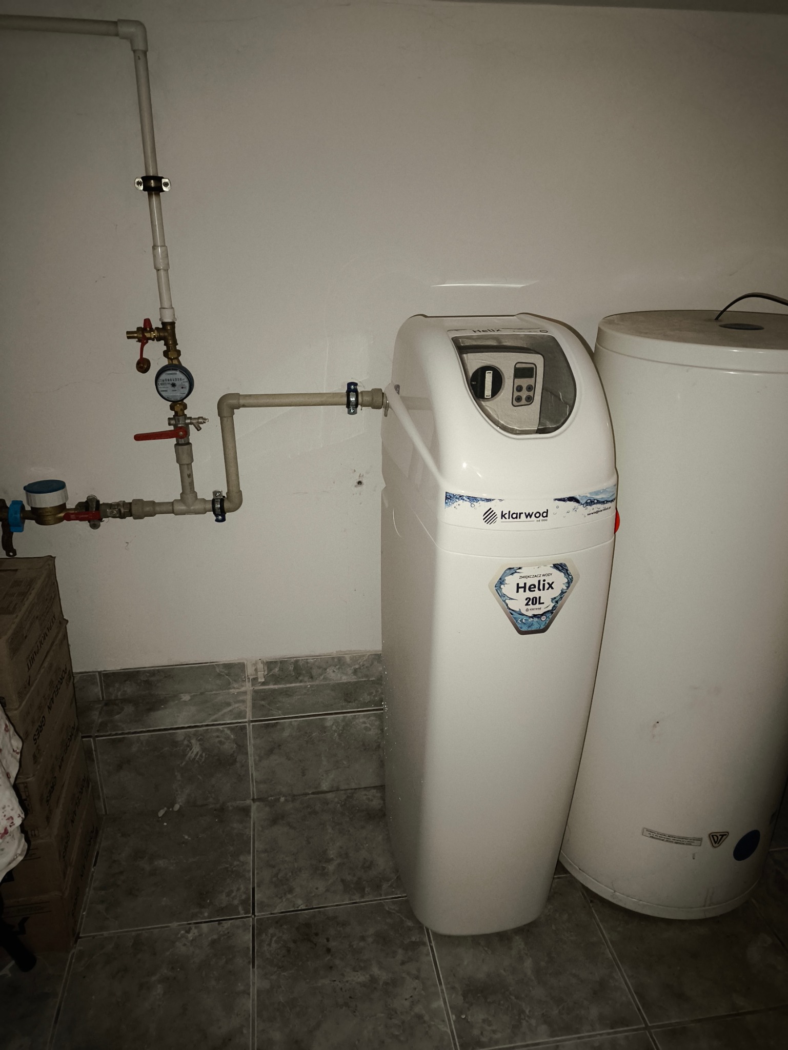 Biały zmiękczacz wody Klarwod Helix 20L, z podłączonymi rurami i zaworami. Obok widoczny biały bojler. Szare płytki na podłodze.