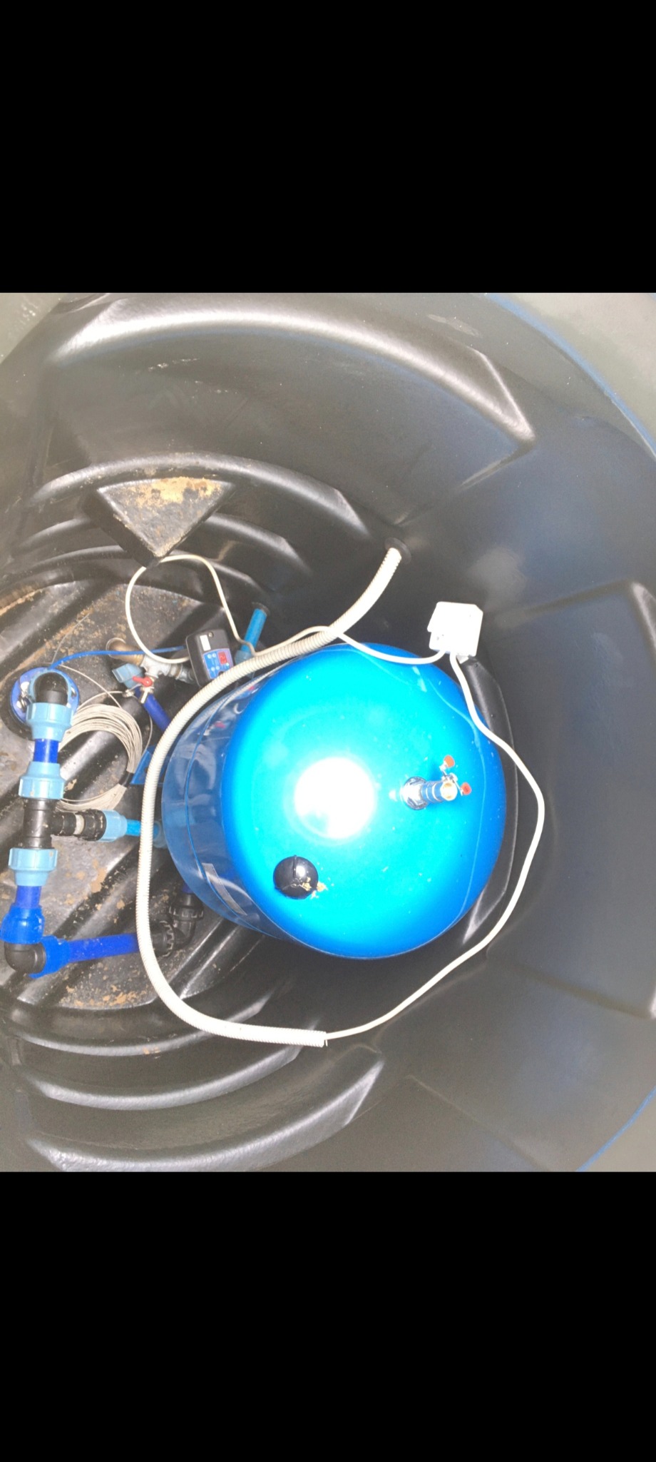 Niebieski hydrofor w czarnym zbiorniku z podłączonymi rurami z tworzywa i przewodami, widok z góry. Instalacja hydrauliczna wewnątrz zbiornika.