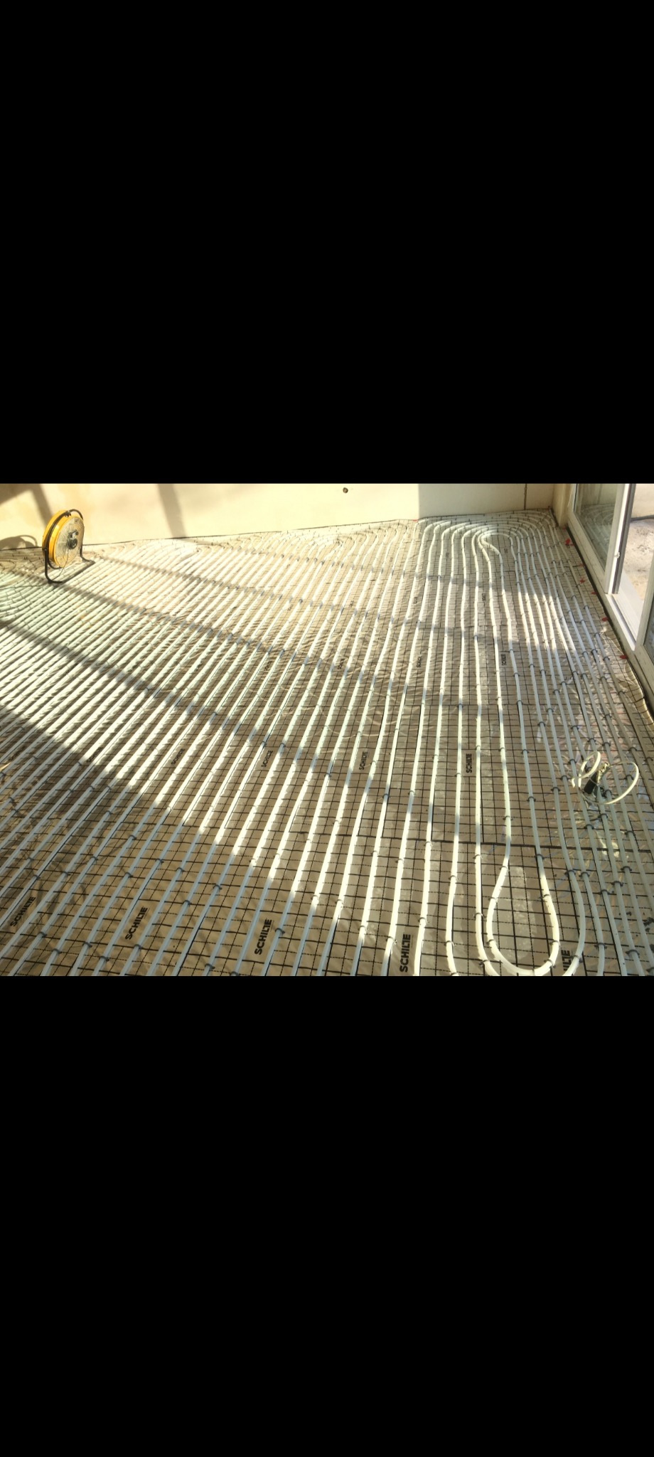 Białe rurki ogrzewania podłogowego ułożone na siatce, z widocznym żółtym wentylatorem i fragmentem okna, oświetlone naturalnym światłem.