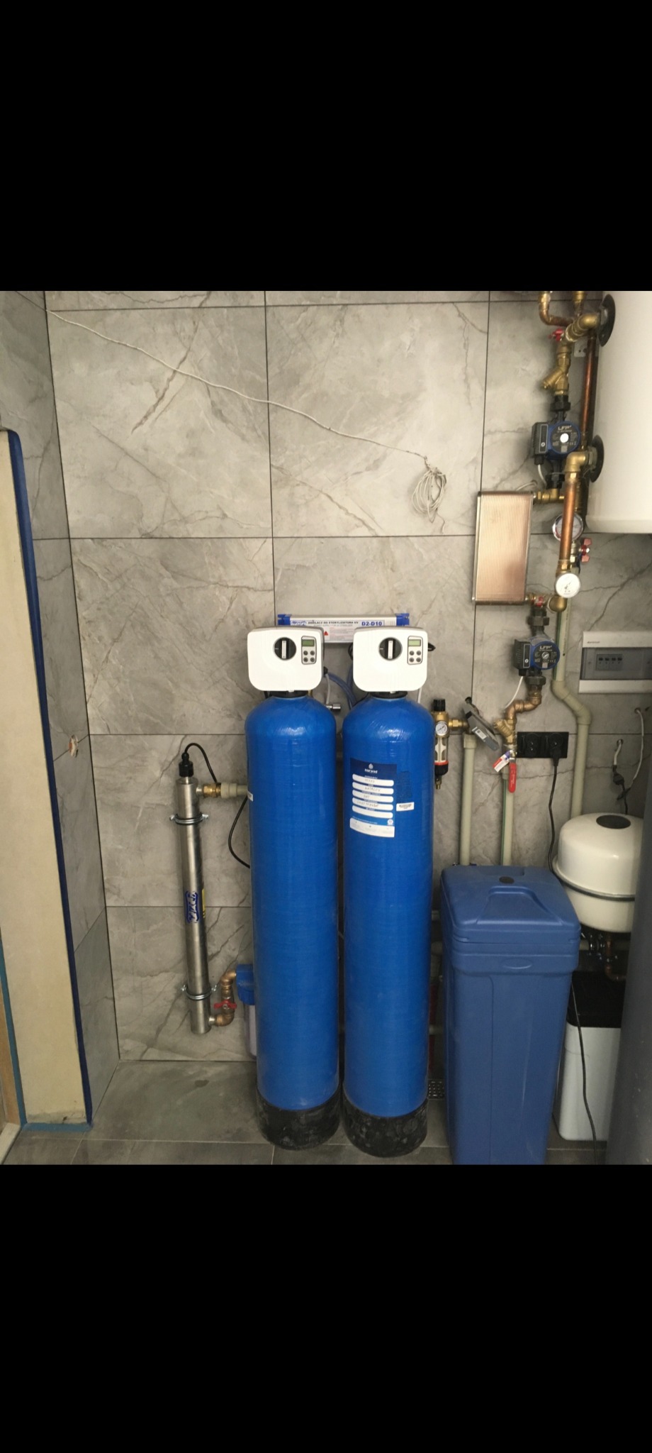 Dwa niebieskie filtry wody z białymi głowicami w pomieszczeniu z szarymi płytkami. Widoczna instalacja wodna z rurami miedzianymi i zbiornik wyrównawczy.