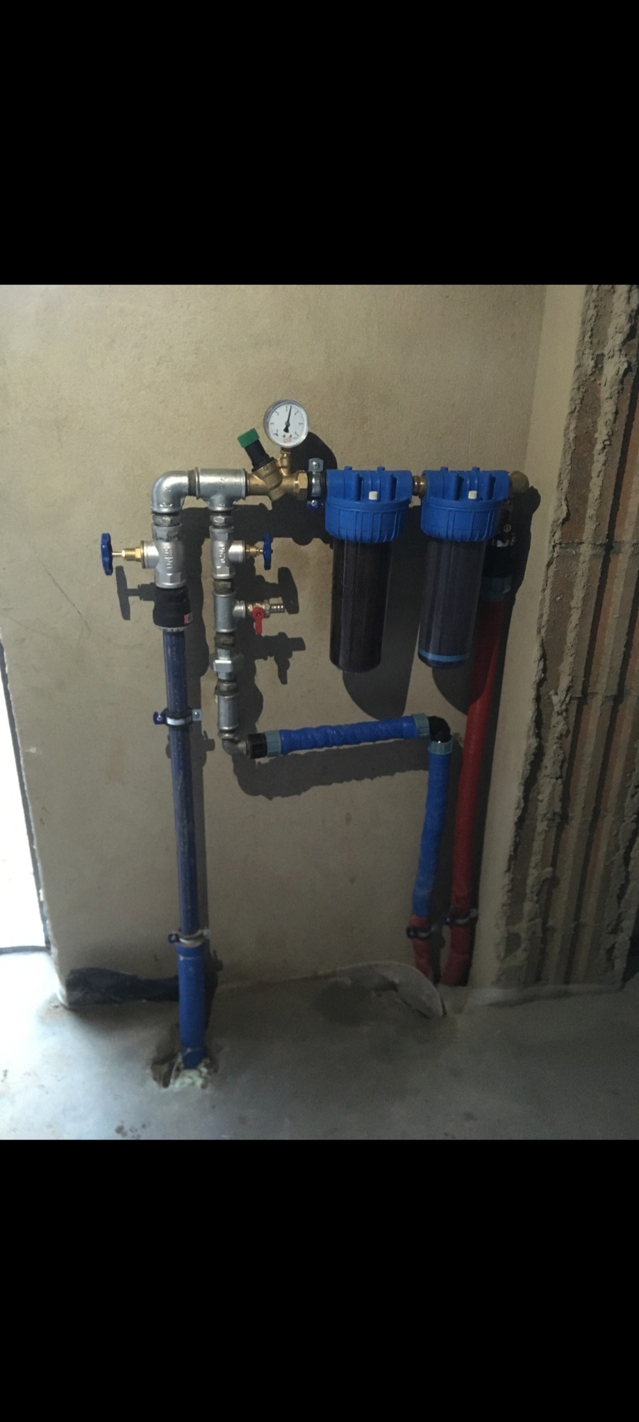 Złożony system filtrów wody z manometrem, rurami z niebieską izolacją i zaworami zamontowany na ścianie w surowym wnętrzu. Widoczne złącza i elementy instalacji.