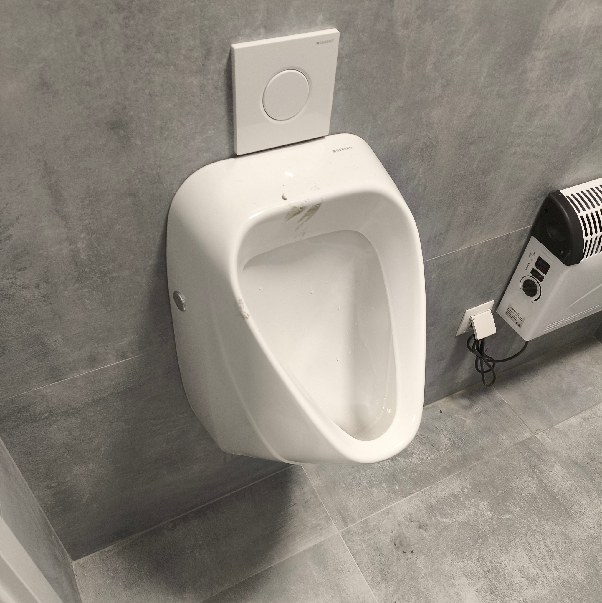 Wiszący, biały pisuar marki Geberit na szarej, betonowej ścianie. Obok grzejnik elektryczny. Widoczne płytki podłogowe w odcieniach szarości.