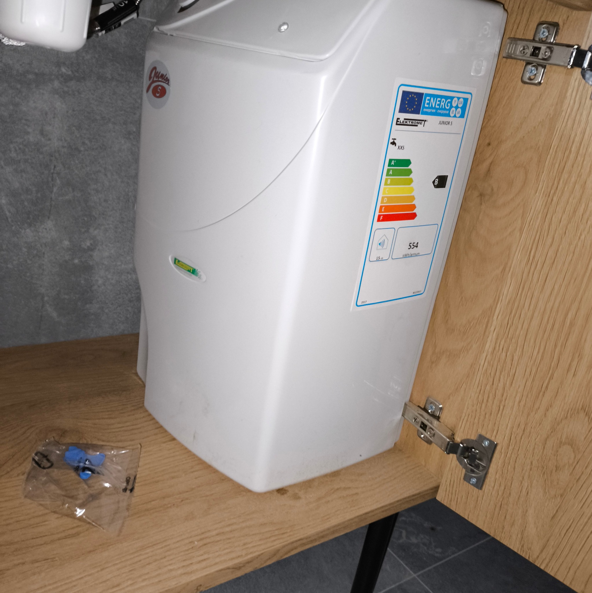 Biały, kompaktowy podgrzewacz wody marki Elektromet Junior S, zamontowany w szafce, z widoczną etykietą energetyczną i akcesoriami w folii obok.