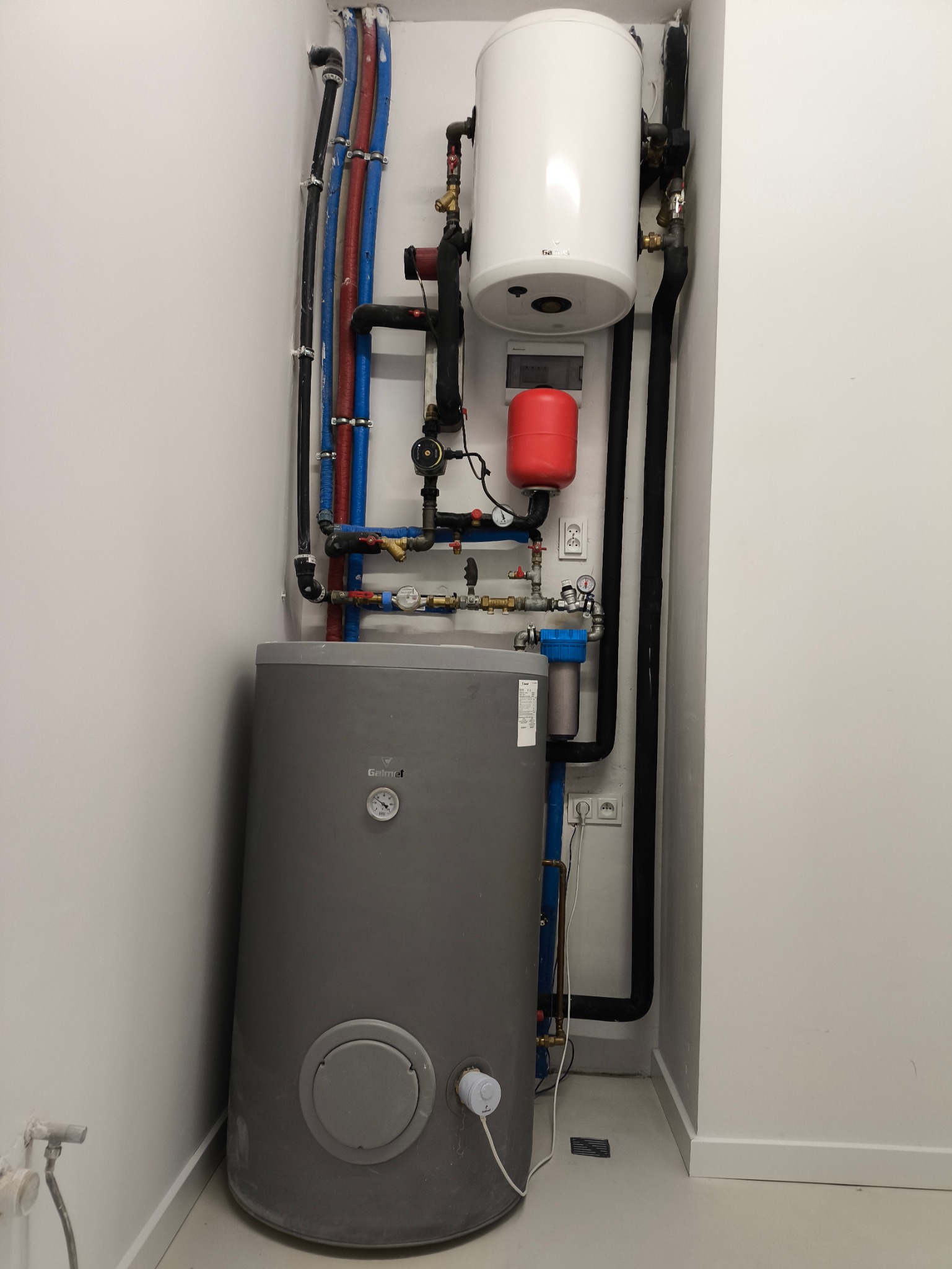 Zestaw instalacji grzewczej z szarym zasobnikiem, białym bojlerem i czerwonym naczyniem wzbiorczym, rurami w kolorach niebieskim, czerwonym i czarnym, na tle białej ściany.