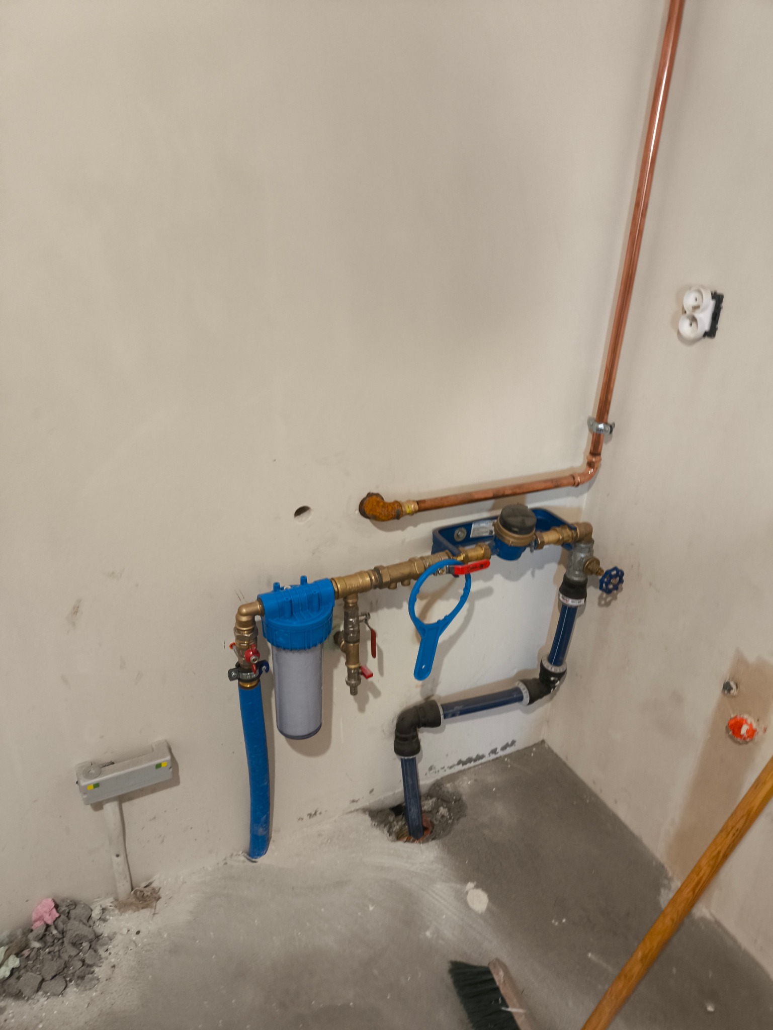 Instalacja wodna z filtrem i licznikiem, rury miedziane i plastikowe, nieotynkowana ściana, widoczne ślady po pracach budowlanych. Elementy instalacji w kolorze miedzianym i niebieskim.