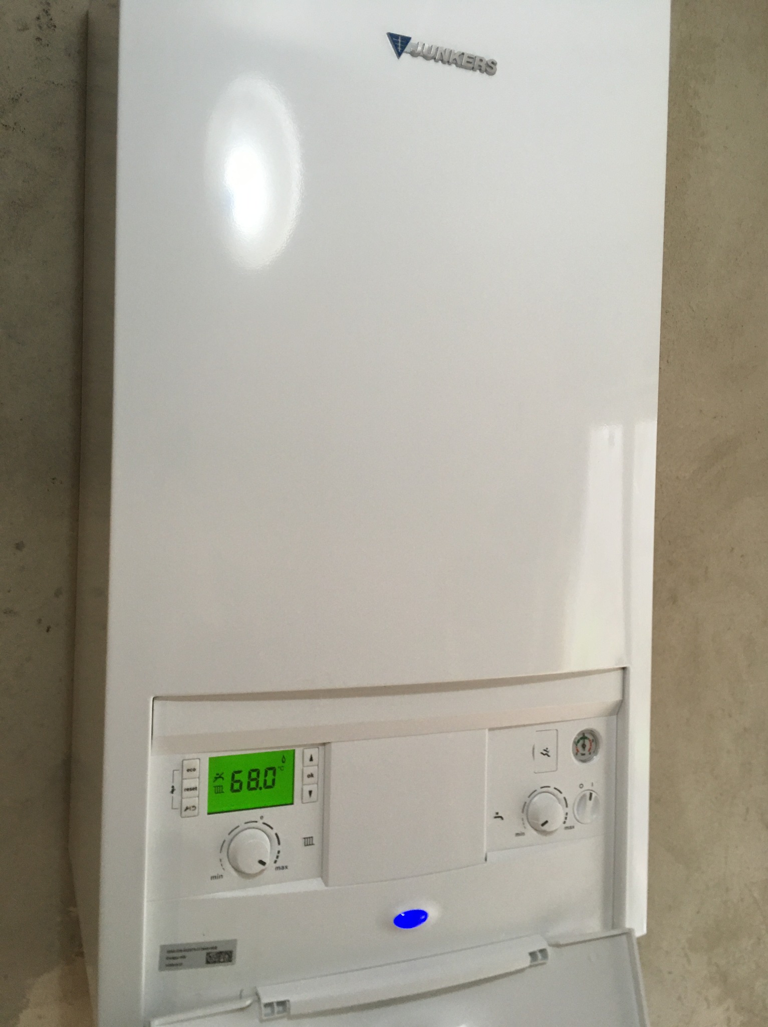 Biały, wiszący piec gazowy Junkers z wyświetlaczem temperatury 68.0°C i regulacją. Widoczny fragment ściany o fakturze betonu.