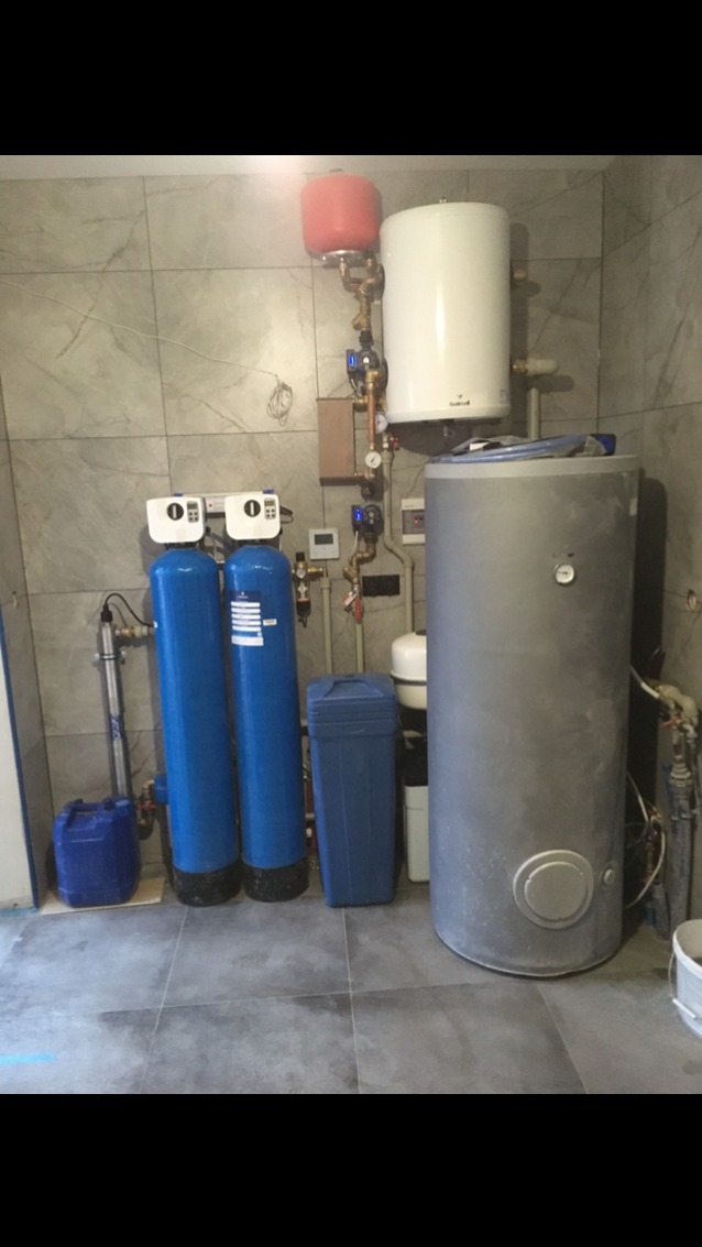 Zestaw instalacji: dwa niebieskie filtry wody, szary bojler, biały zasobnik i czerwony zbiornik wyrównawczy na tle szarych płytek. Elementy połączone miedzianymi rurami.