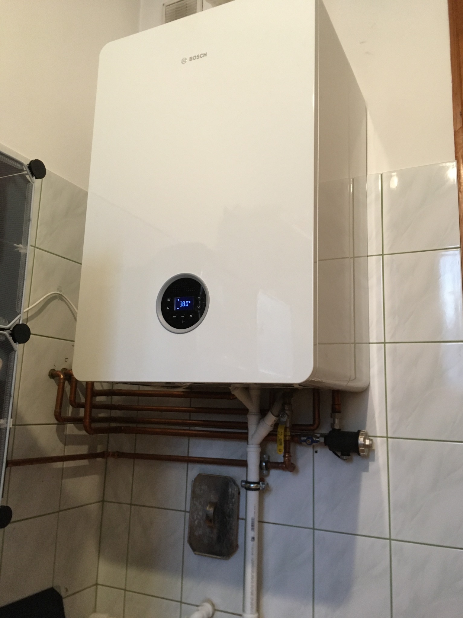 Biały, wiszący piec gazowy marki Bosch z wyświetlaczem 38.0°C, zamontowany na ścianie wyłożonej kafelkami. Widoczne miedziane rury i elementy instalacji.