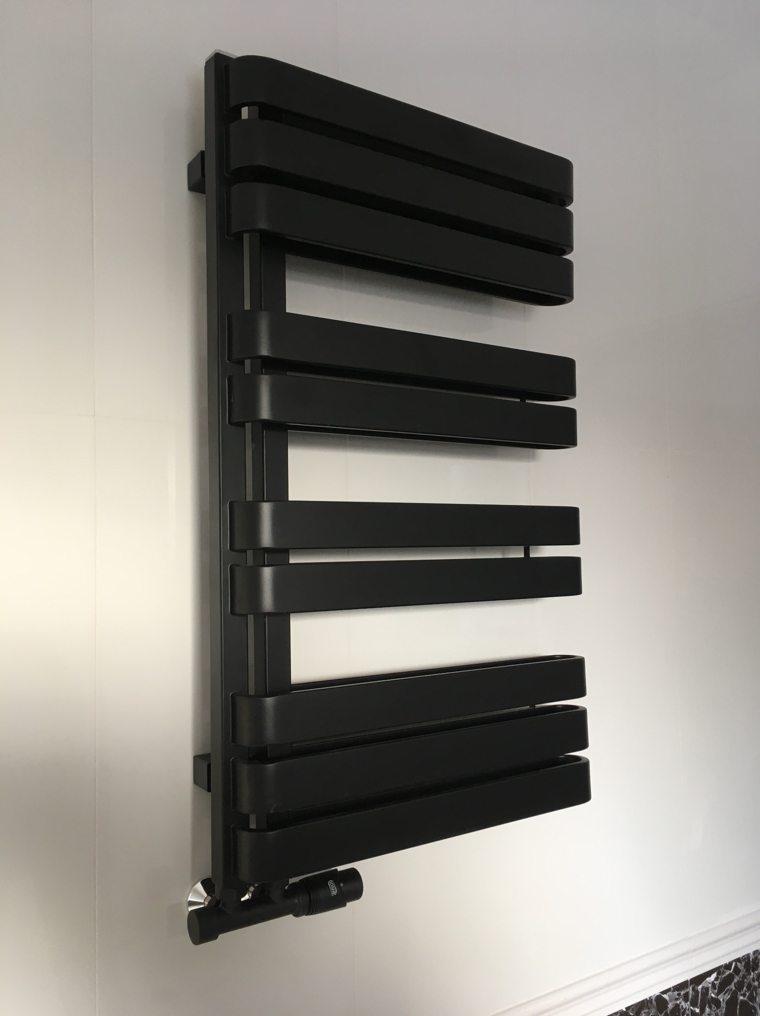 Czarny, designerski grzejnik łazienkowy, zamontowany na ścianie z białych płytek. Widoczny zawór termostatyczny. Minimalistyczny styl.