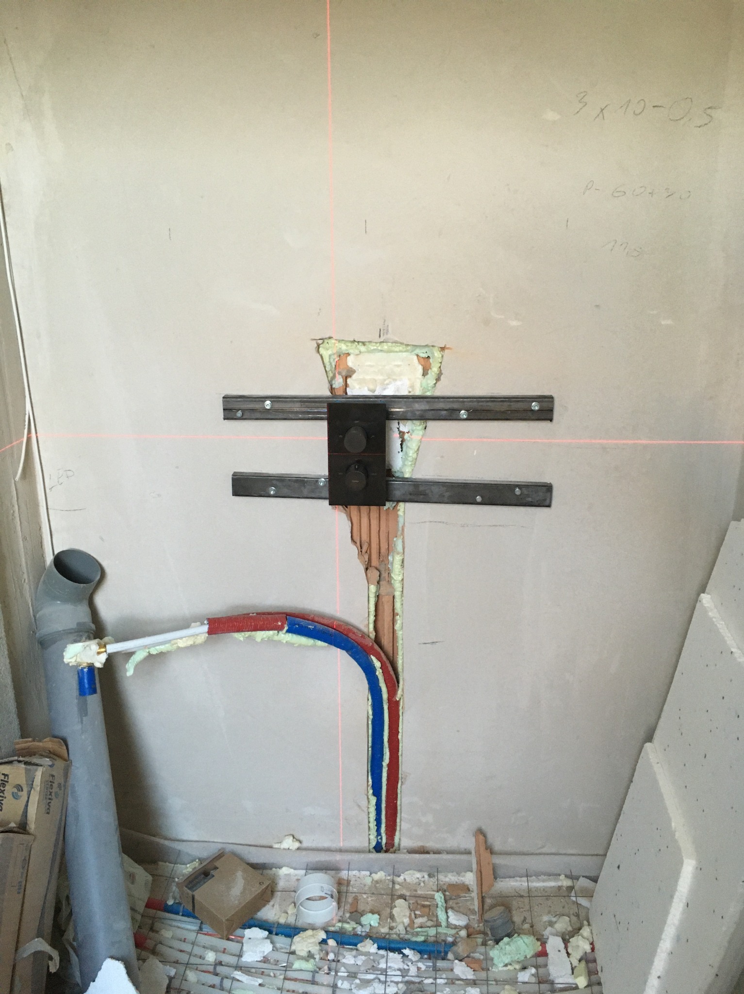 Ściana z wyciętym otworem pod instalację, widoczne rury w izolacji (czerwona i niebieska), metalowe uchwyty montażowe, laserowy poziom, fragment podłogi z instalacją.