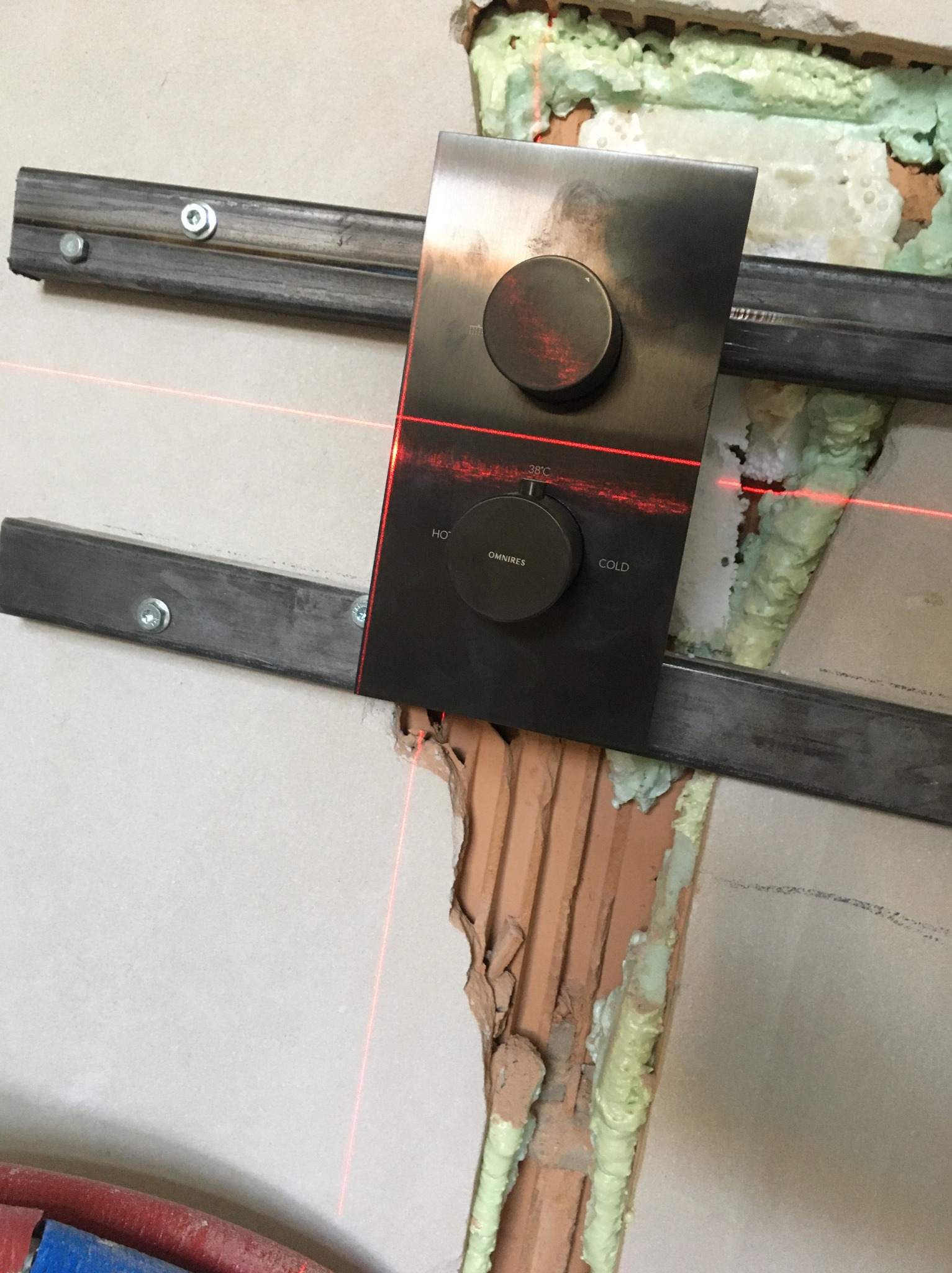 Montaż panelu sterowania prysznicem Omnires z użyciem poziomicy laserowej, widoczne metalowe szyny montażowe i pianka uszczelniająca w ścianie.