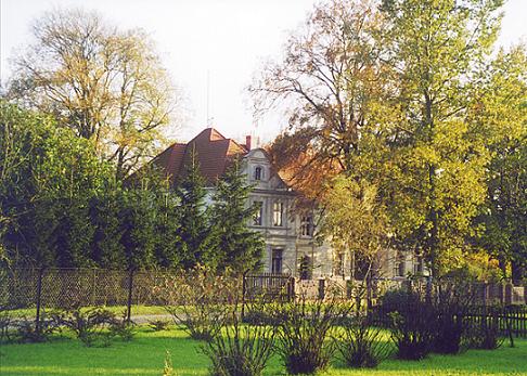 Dwór w Boduszewie z czerwonym dachem, otoczony zielenią drzew i krzewów, widoczny zza niskiego ogrodzenia.