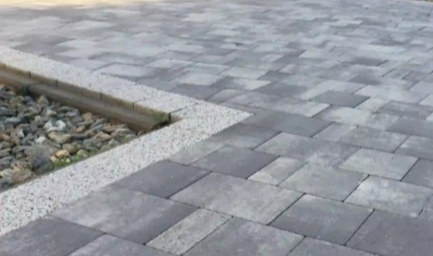 Szara kostka brukowa ułożona w regularny wzór, obrzeże z jasnego grysu i kamieni ozdobnych, widok z góry, lekki cień.