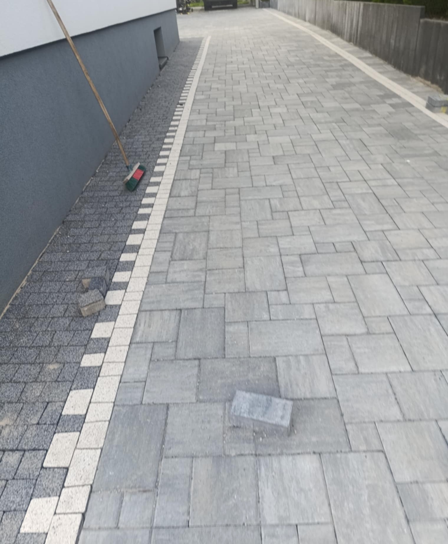 Ułożona kostka brukowa w szaro-białej kolorystyce, widoczny fragment ściany budynku i szczotka. Częściowo widoczne niezakończone prace.