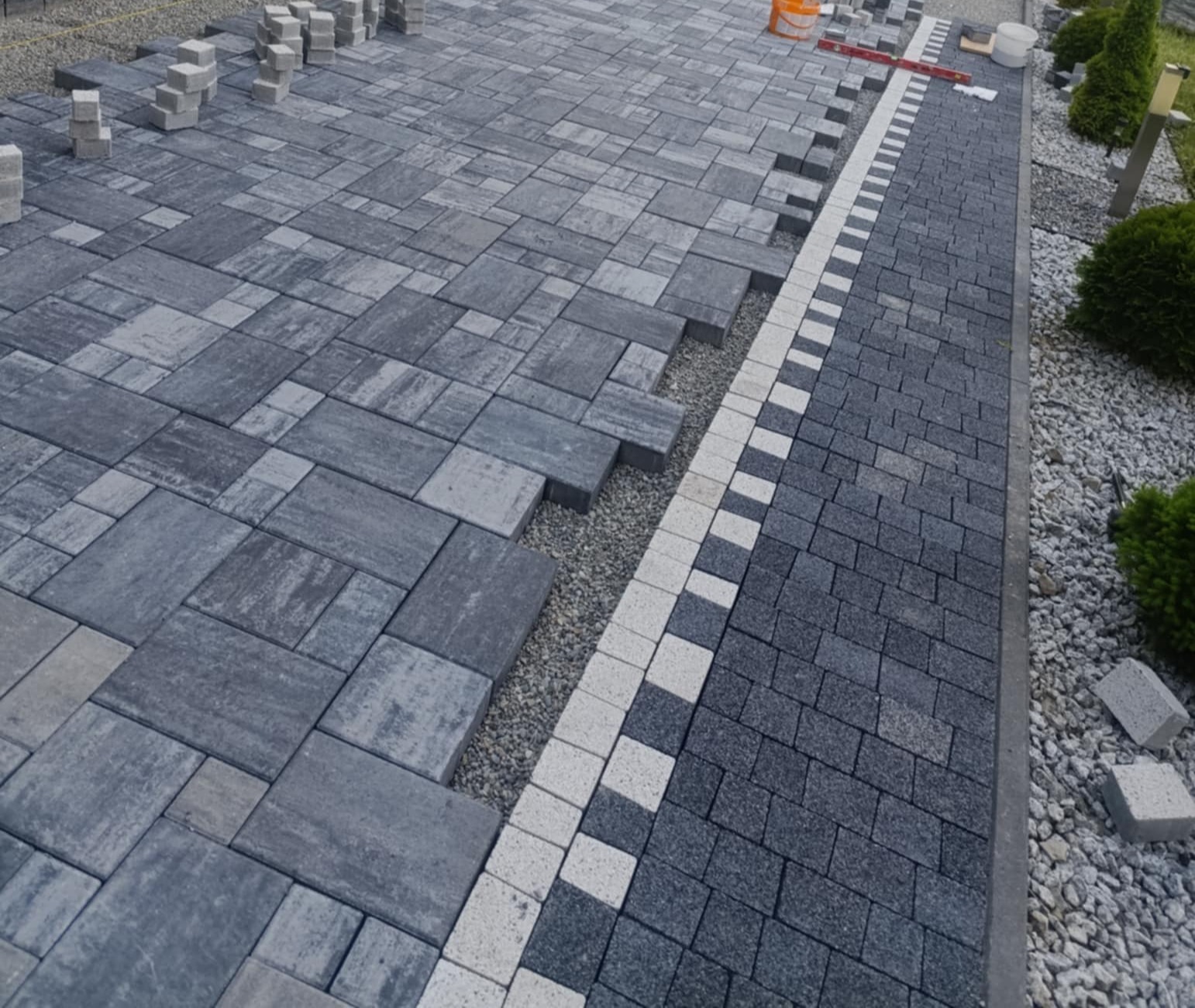 Fragment układania szarej kostki brukowej z białym obramowaniem; widoczne przygotowane podłoże i ułożone fragmenty nawierzchni.