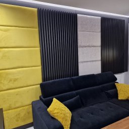 Meblowy Design Arkadiusz Grzywna - Panel ścienny z żółtymi i beżowymi poduszkami obok czarnych pionowych listew dekoracyjnych, uzupełniony czarną sofą narożną z żółtymi poduszkami i lampą podłogową