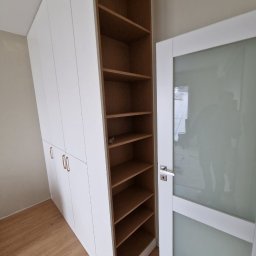 Meblowy Design Arkadiusz Grzywna - Biała szafa z frontami na wysoki połysk, zintegrowana z regałem z drewnianymi półkami, obok drzwi z matową szybą.