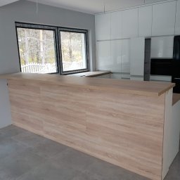 Meblowy Design Arkadiusz Grzywna - Jasna kuchnia z drewnianym blatem wyspowym i białymi szafkami, widok na zimowy krajobraz przez okno.