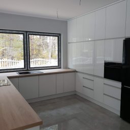 Meblowy Design Arkadiusz Grzywna - Jasna kuchnia z białymi, błyszczącymi frontami, drewnianym blatem i dużym oknem z widokiem na ogród, czarna lodówka Samsung po prawej stronie.