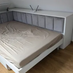 Białe łóżko na wymiar z szarym, tapicerowanym wezgłowiem w pionowe panele, z półką na górze, na której stoi router WiFi. Łóżko znajduje się na poddaszu.
