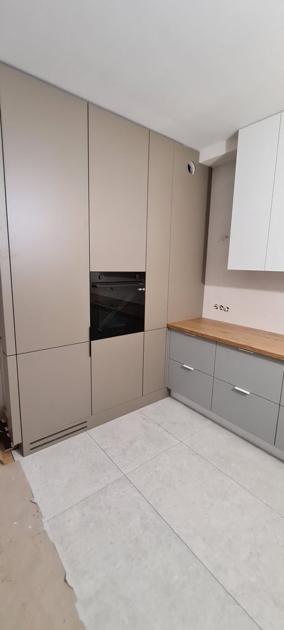 Nowoczesna zabudowa kuchenna z beżowymi szafkami, czarnym piekarnikiem i szarymi dolnymi szafkami z drewnianym blatem, na jasnych płytkach podłogowych.