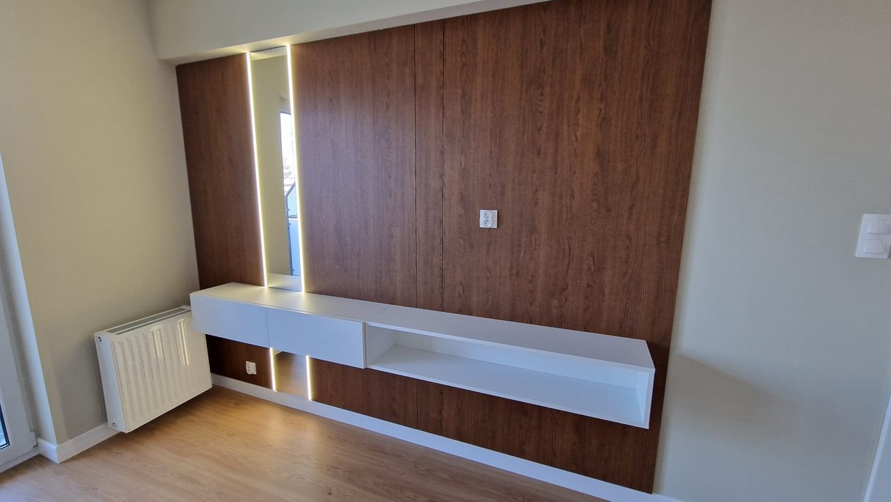 Drewniana ściana z wbudowanym oświetleniem LED i białą szafką RTV z szufladą, uzupełniona grzejnikiem na ścianie po lewej stronie.
