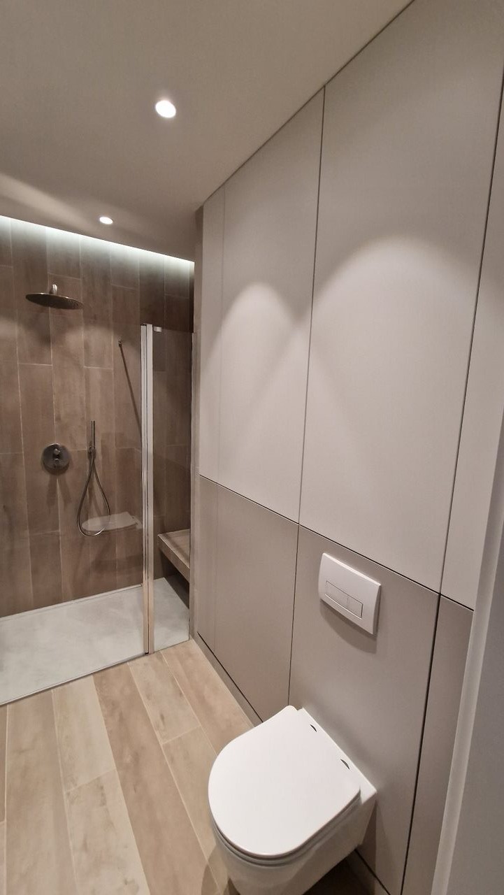 Nowoczesna łazienka z zabudową meblową w odcieniach szarości, kabina prysznicowa typu walk-in z deszczownicą, wisząca miska WC i podłoga imitująca drewno.