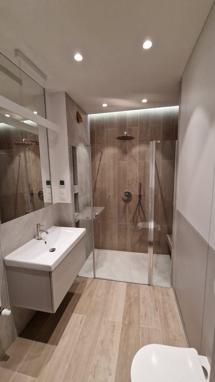 Nowoczesna łazienka z kabiną prysznicową typu walk-in, umywalką nablatową z szafką w kolorze beżowym i podłogą wyłożoną płytkami imitującymi drewno.