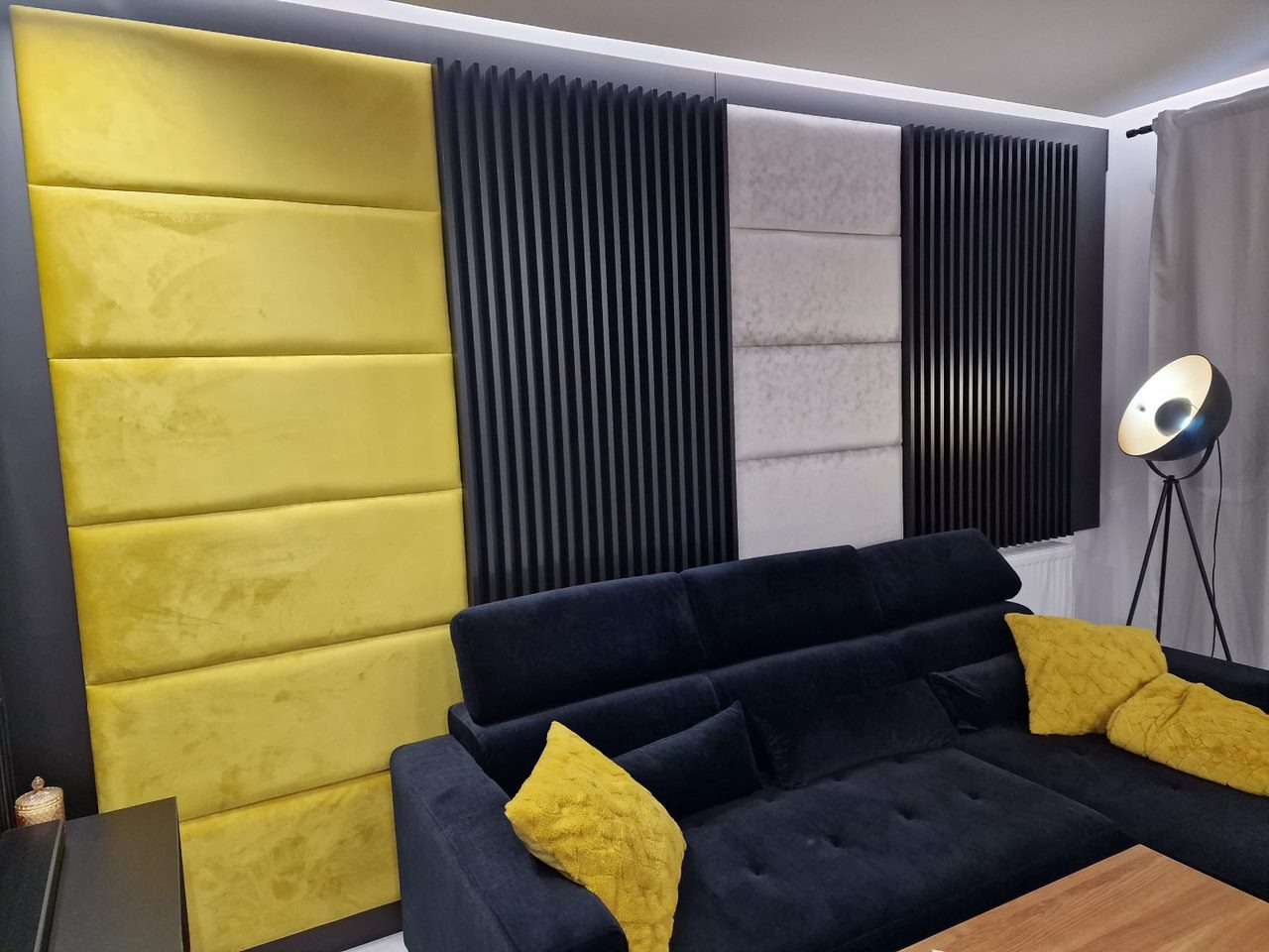 Panel ścienny z żółtymi i beżowymi poduszkami obok czarnych pionowych listew dekoracyjnych, uzupełniony czarną sofą narożną z żółtymi poduszkami i lampą podłogową