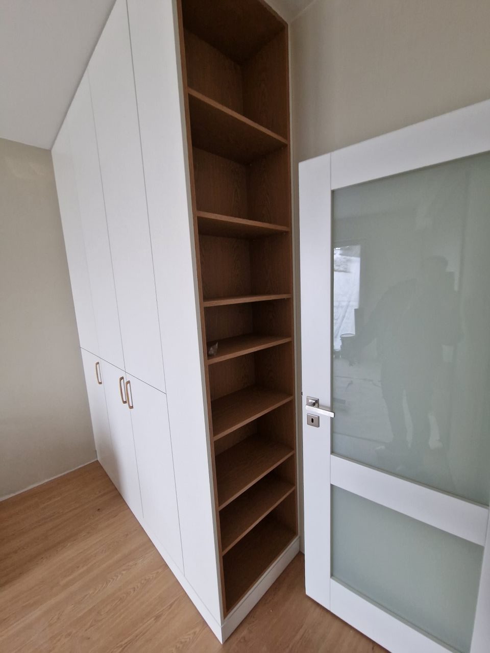 Biała szafa z frontami na wysoki połysk, zintegrowana z regałem z drewnianymi półkami, obok drzwi z matową szybą.