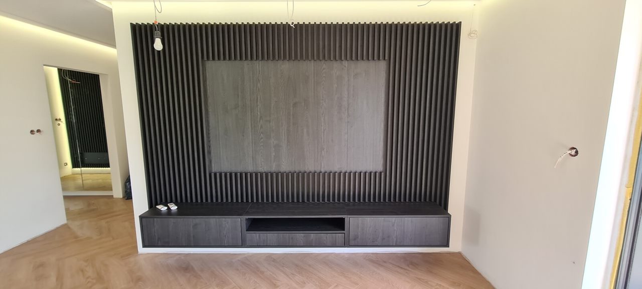 Ciemnoszara zabudowa ściany z lamelami i szafką RTV w minimalistycznym salonie, widoczne kable oświetleniowe i niedokończone wykończenie.