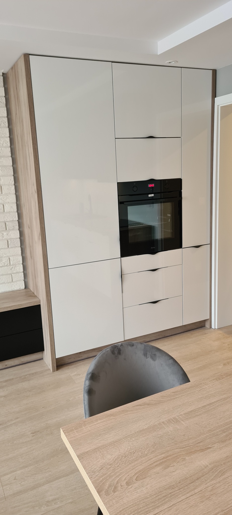 Wysoka zabudowa kuchenna z białymi, lakierowanymi frontami, wbudowanym piekarnikiem i dyskretnymi uchwytami, z boku wykończenie imitujące drewno, widoczny fragment blatu stołu i krzesła.