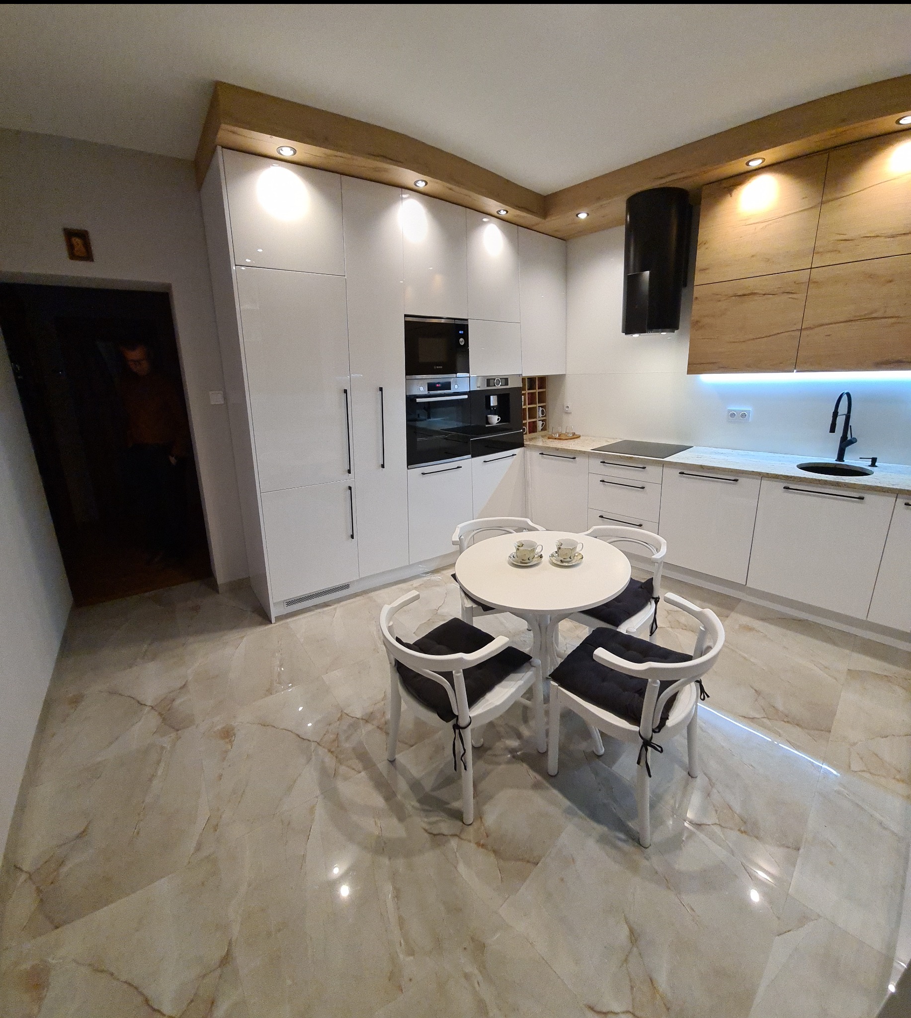 Nowoczesna kuchnia z białymi, lakierowanymi frontami, drewnianym akcentem na górze szafek, czarnym okapem i okrągłym stołem z krzesłami na marmurowej podłodze.