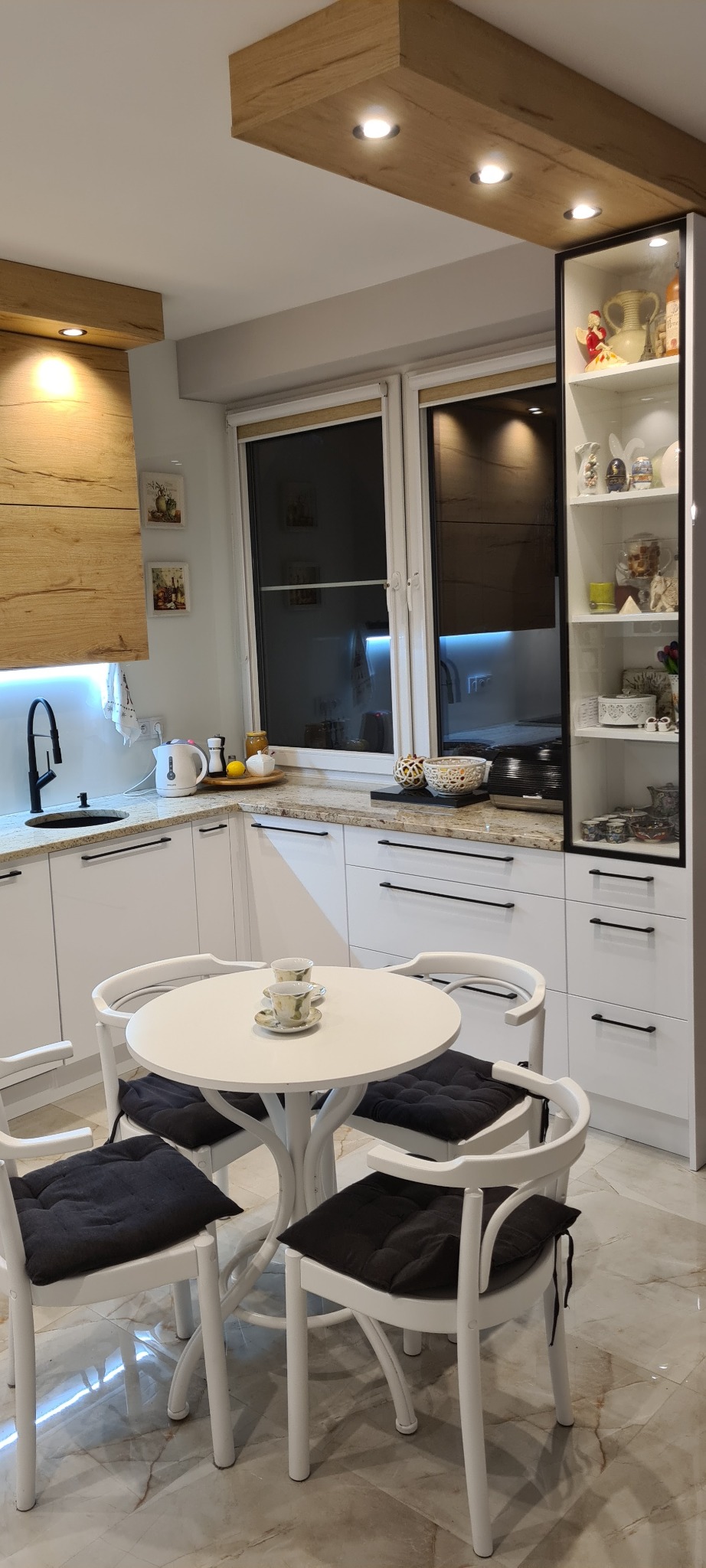Jasna kuchnia z białymi szafkami, granitowym blatem, czarną armaturą i okrągłym stołem z filiżankami, oświetlona drewnianymi belkami sufitowymi z punktowymi lampami.