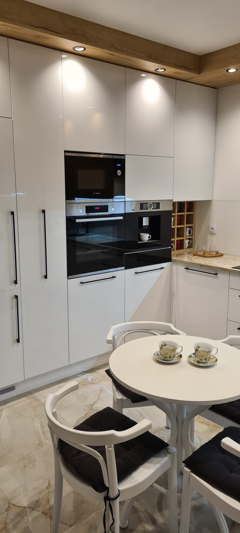 Jasna kuchnia z białymi, lakierowanymi frontami, zabudowanymi urządzeniami marki Bosch i okrągłym stołem z filiżankami.